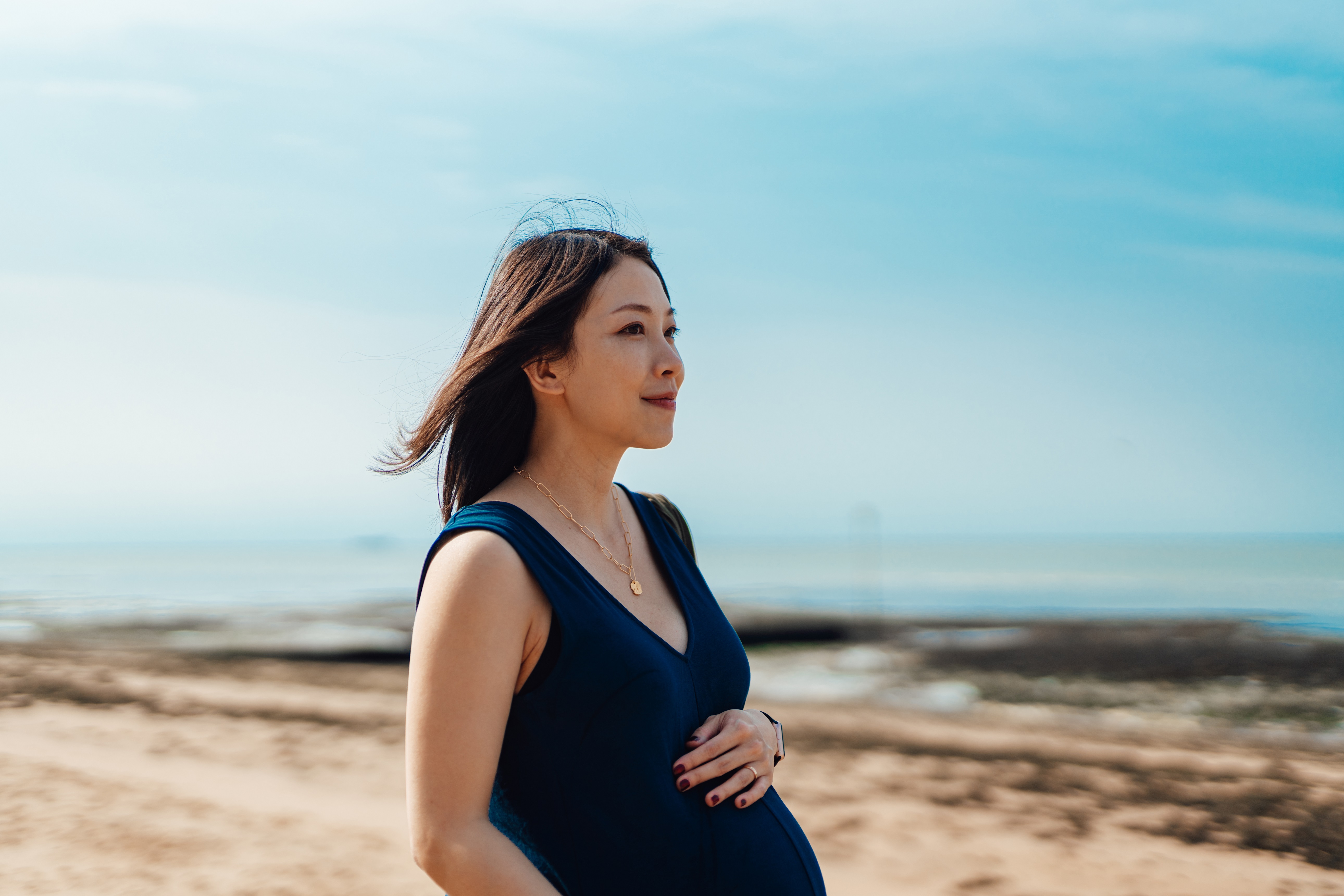 Rear view of an Asian young pregnant woman having a walk by the beach. Touching her bump. Welcome a new life to be born. By the window. Healthy and active pregnancy lifestyle. Third trimester.